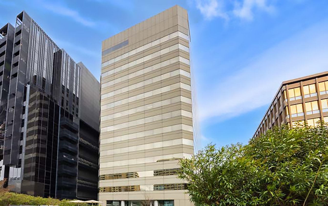 Tall modern office buildings in an urban area with a blue sky and green trees in the foreground.