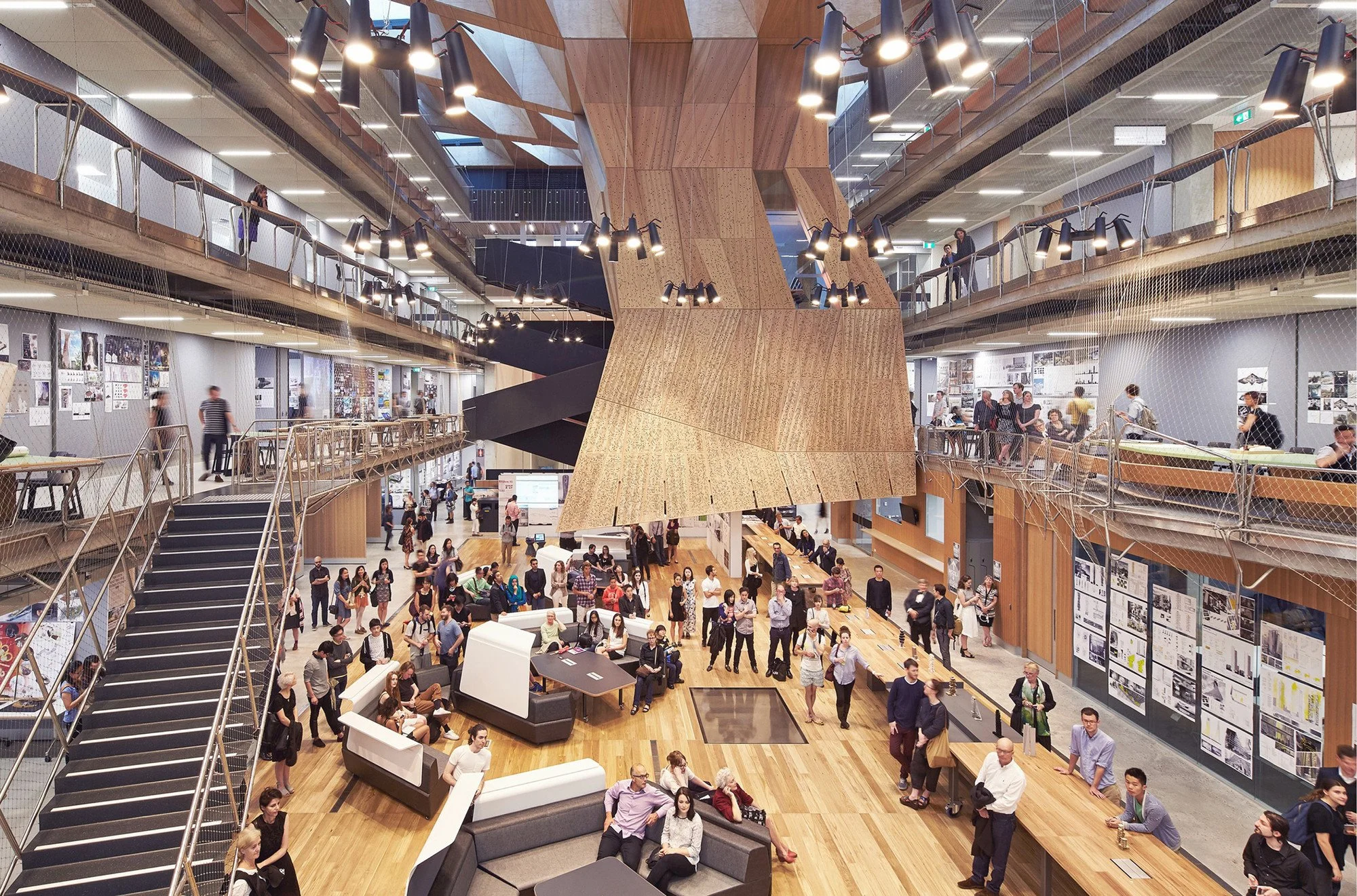 A spacious multi-level indoor space filled with people. There are wooden floors, modern lighting, and a large wooden architectural feature in the center. Multiple people are walking, sitting, and talking, with some looking at displays or working at t