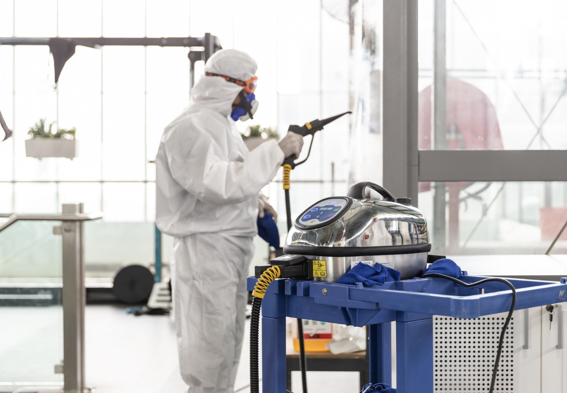 A person dressed in protective medical gear, including a white suit, gloves, and goggles, is cleaning or disinfecting a surface with a spray device in a bright indoor space, possibly an airport or hospital.