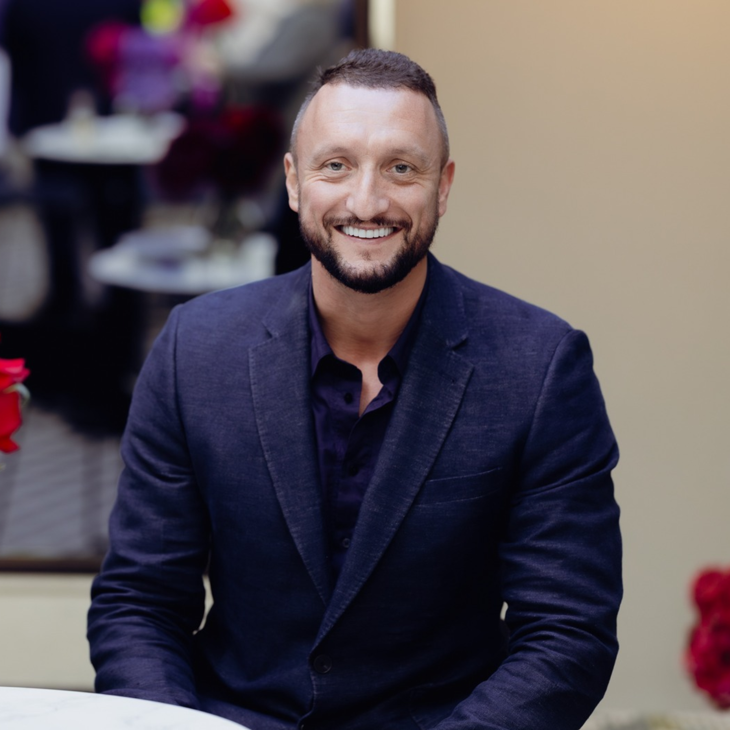 A man with short dark hair, a beard, and a bright smile, wearing a dark blazer over a dark shirt, sitting indoors with a blurred background.