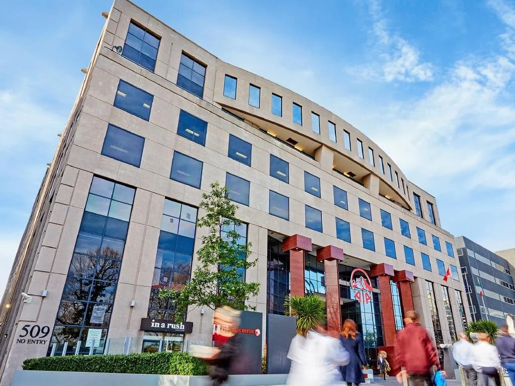 Modern multi-story office building with a glass facade and the logo of AI in front, people walking on the sidewalk, and a clear blue sky.