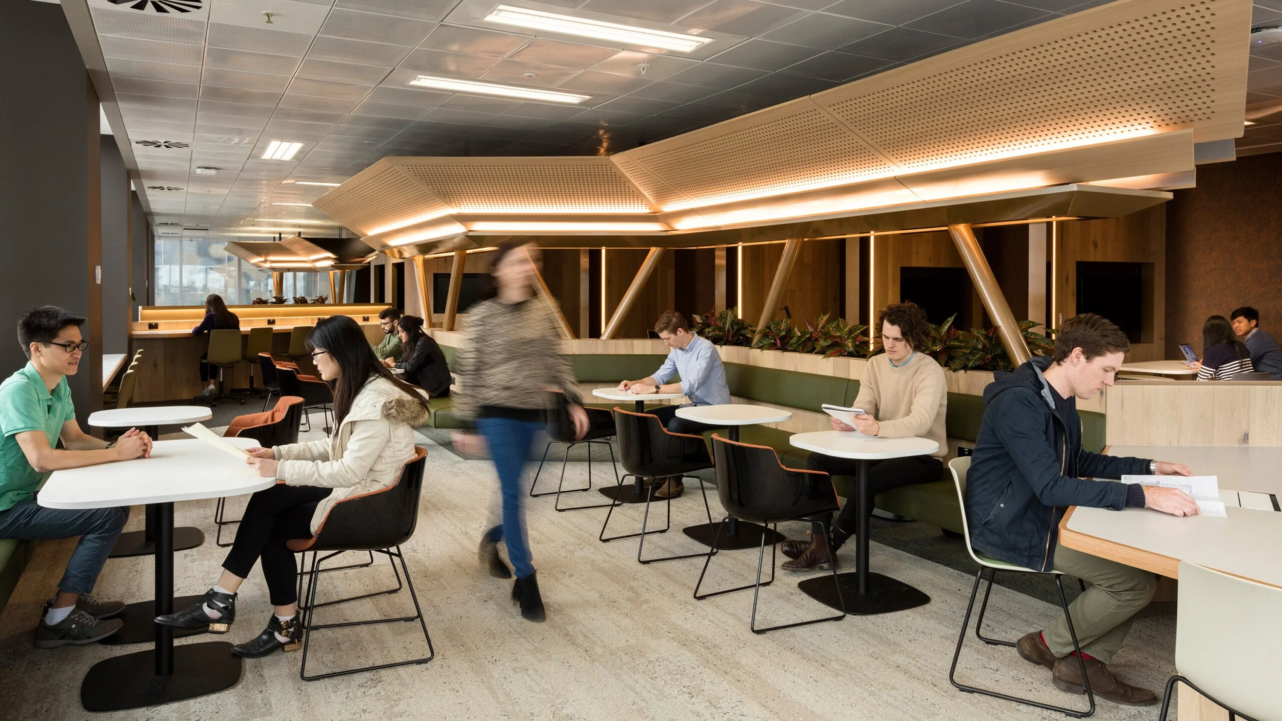People sitting at tables in a modern, well-lit indoor space, some reading or using electronic devices, with a woman walking through the scene.