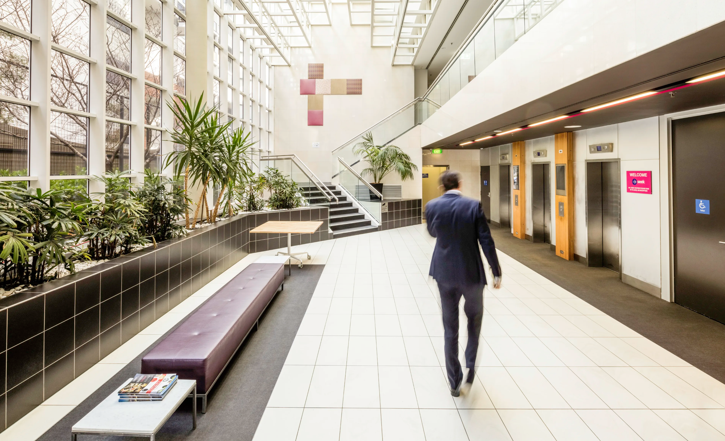 Interior of a modern building lobby with large windows, plants, a staircase, and an elevator area with a man walking.