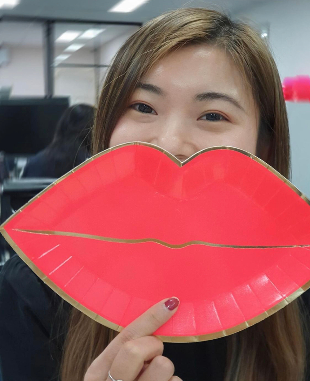 Woman holding a large red lips-shaped sign in front of her face, only her eyes visible, in an office setting.