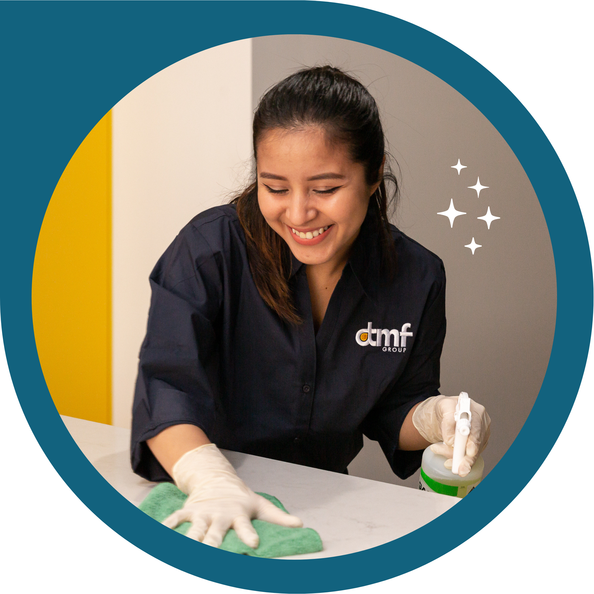 A woman wearing gloves and a black shirt with a company logo, smiling while cleaning a surface with a spray bottle and cloth.