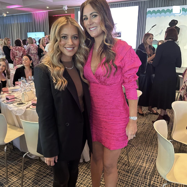 Two women standing together at a social event, smiling, with tables set for a meal in the background.