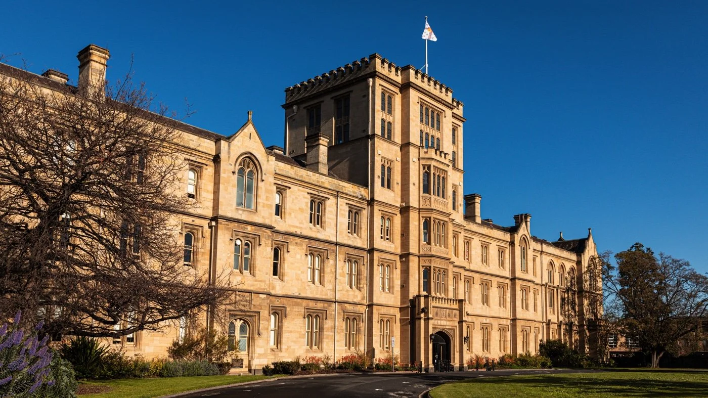 A historic castle-like building made of tan stone with numerous windows, a central tower topped with a flagpole and a flag, surrounded by landscaped grass and trees under a clear blue sky.
