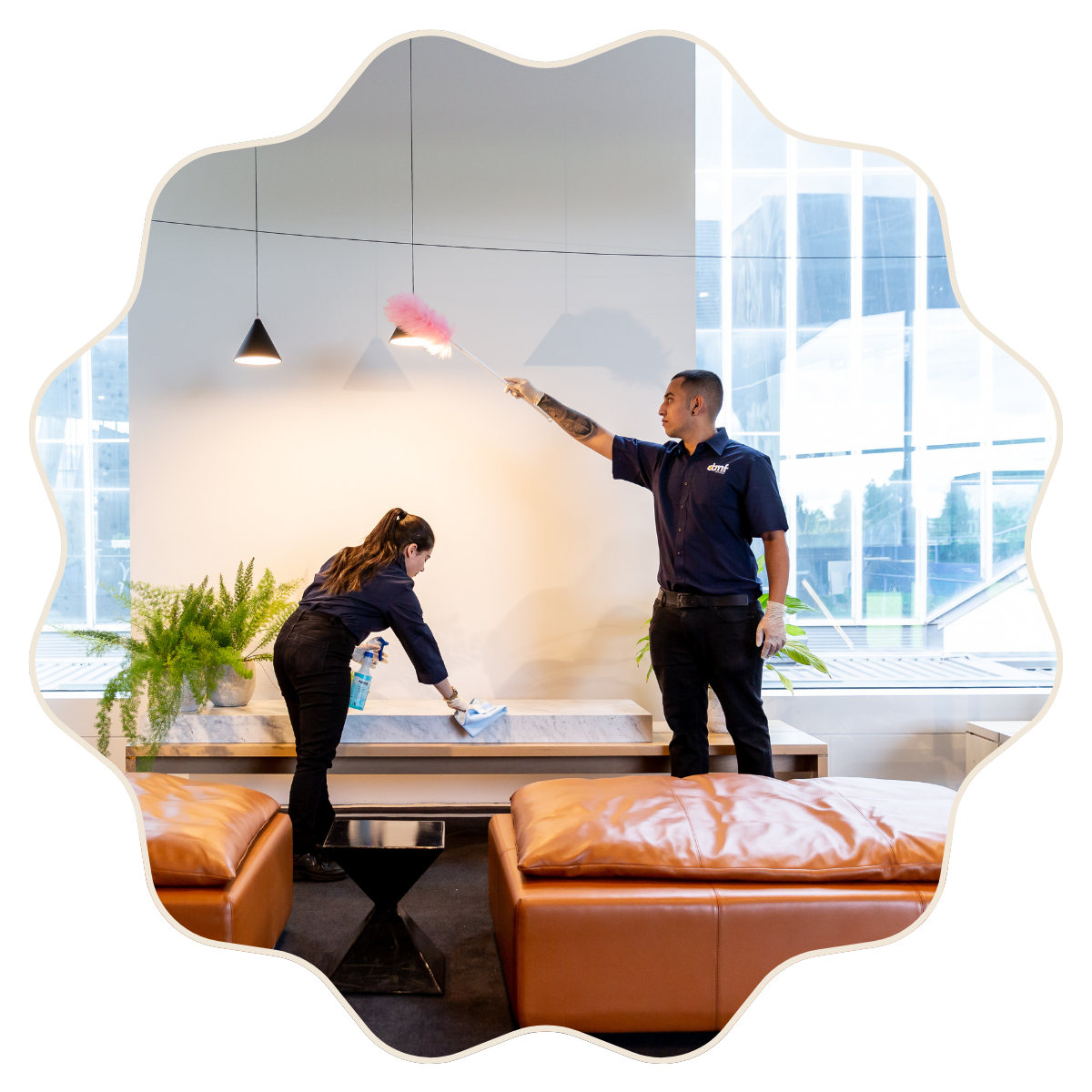 Two hotel staff members cleaning a lobby area with a spray bottle and a cloth, one is spraying the ceiling light while the other is wiping the marble surface.