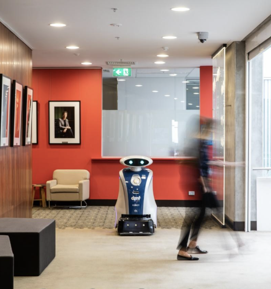 An indoor lobby with a small robot standing in front of a red wall, a white armchair, framed pictures on the wall, and a person walking by blurred due to motion.