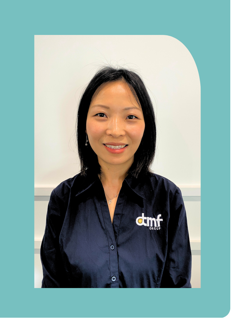 A woman with shoulder-length black hair smiling, wearing a navy blue button-up shirt with a logo that reads 'dmf GROUP', standing against a plain white wall.