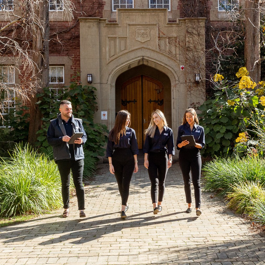 Four people, three women and one man, walking and talking on a brick pathway in front of an old brick building with a wooden door. The women are holding tablets, and the man is holding a tablet. The building has a stone archway with the year 1929 inscribed above the door. There is greenery and trees surrounding the pathway.