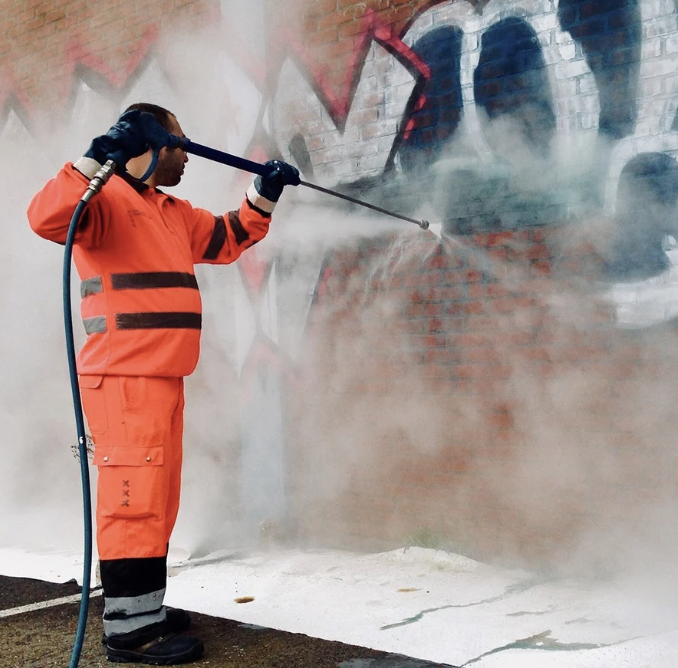 A worker in orange safety gear pressure washing a graffiti-covered brick wall, creating steam and spray.