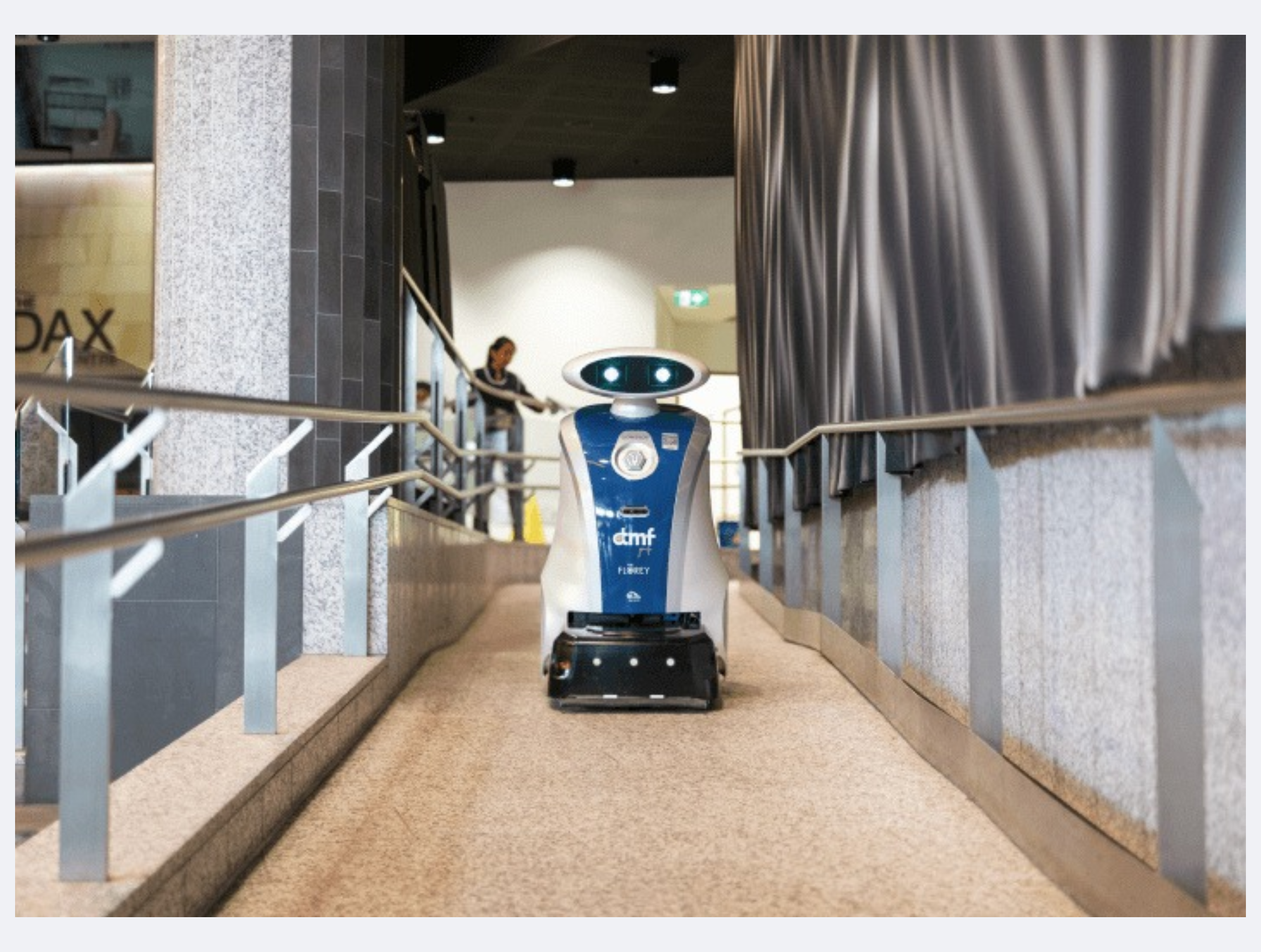 A robot with a blue and white body, two glowing eyes on its head, and the words 'dmf' and 'FLEX' on its chest, moving along a corridor with metal railings on both sides in an indoor building.