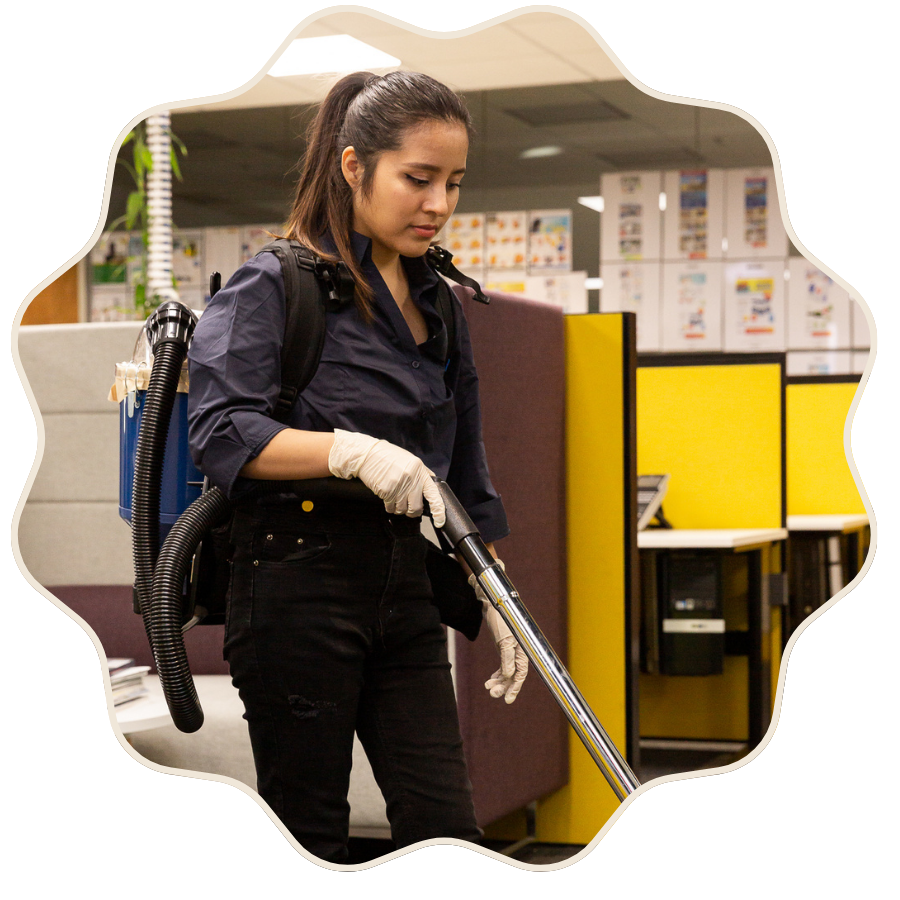 A woman wearing gloves and a backpack vacuuming office cubicles in an indoor workspace.