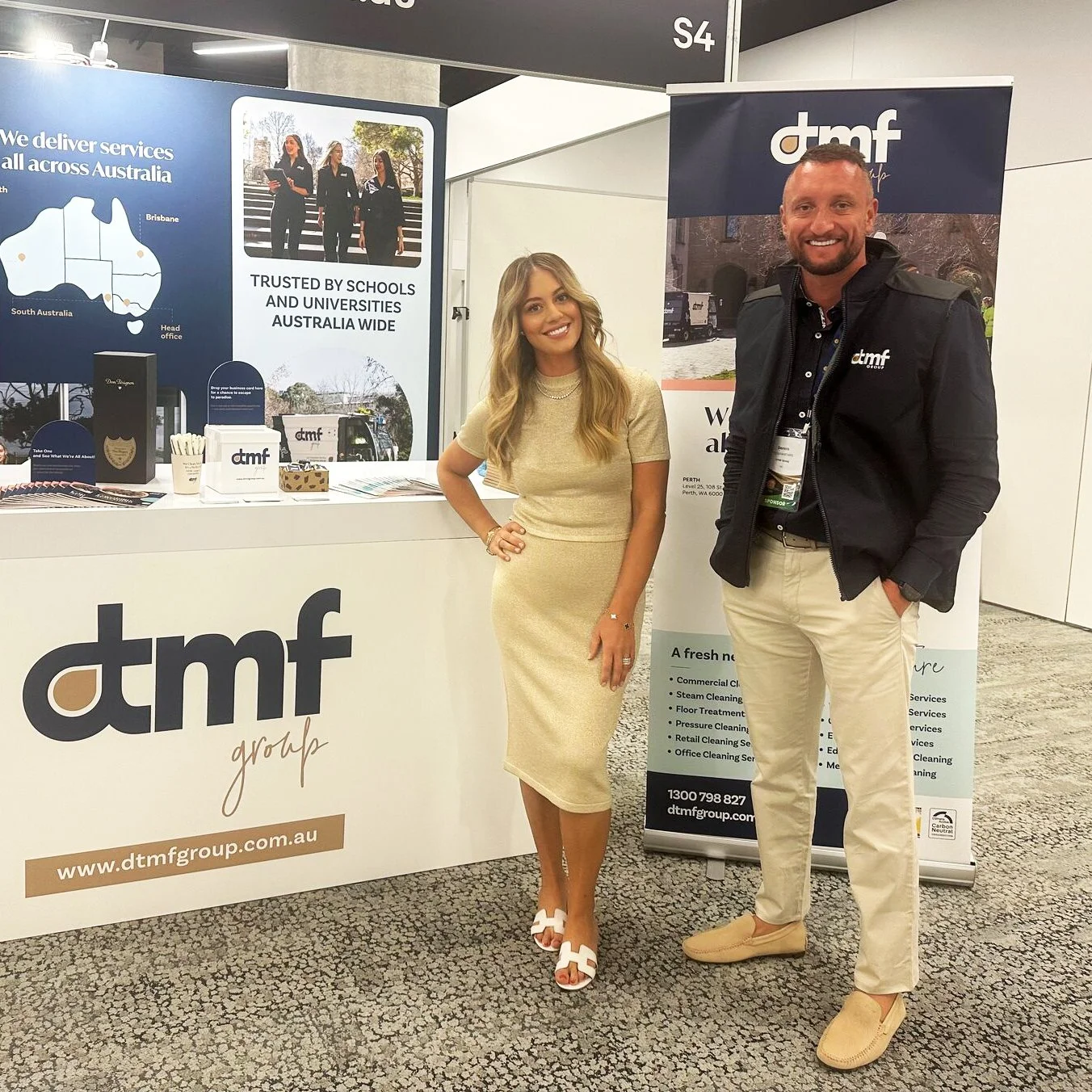 A man and a woman standing together at a booth for dmtf, a cleaning service company, at an event. The woman is wearing a beige dress and white heels, and the man is wearing a black jacket, beige pants, and beige shoes. The booth features banners and marketing materials for dmtf, including a map of Australia and the company's logo.