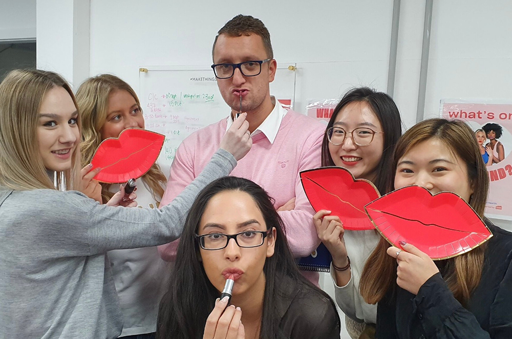 Group of young women and a man posing with large red lipstick-shaped props, and two women applying lipstick, in a classroom or office setting.