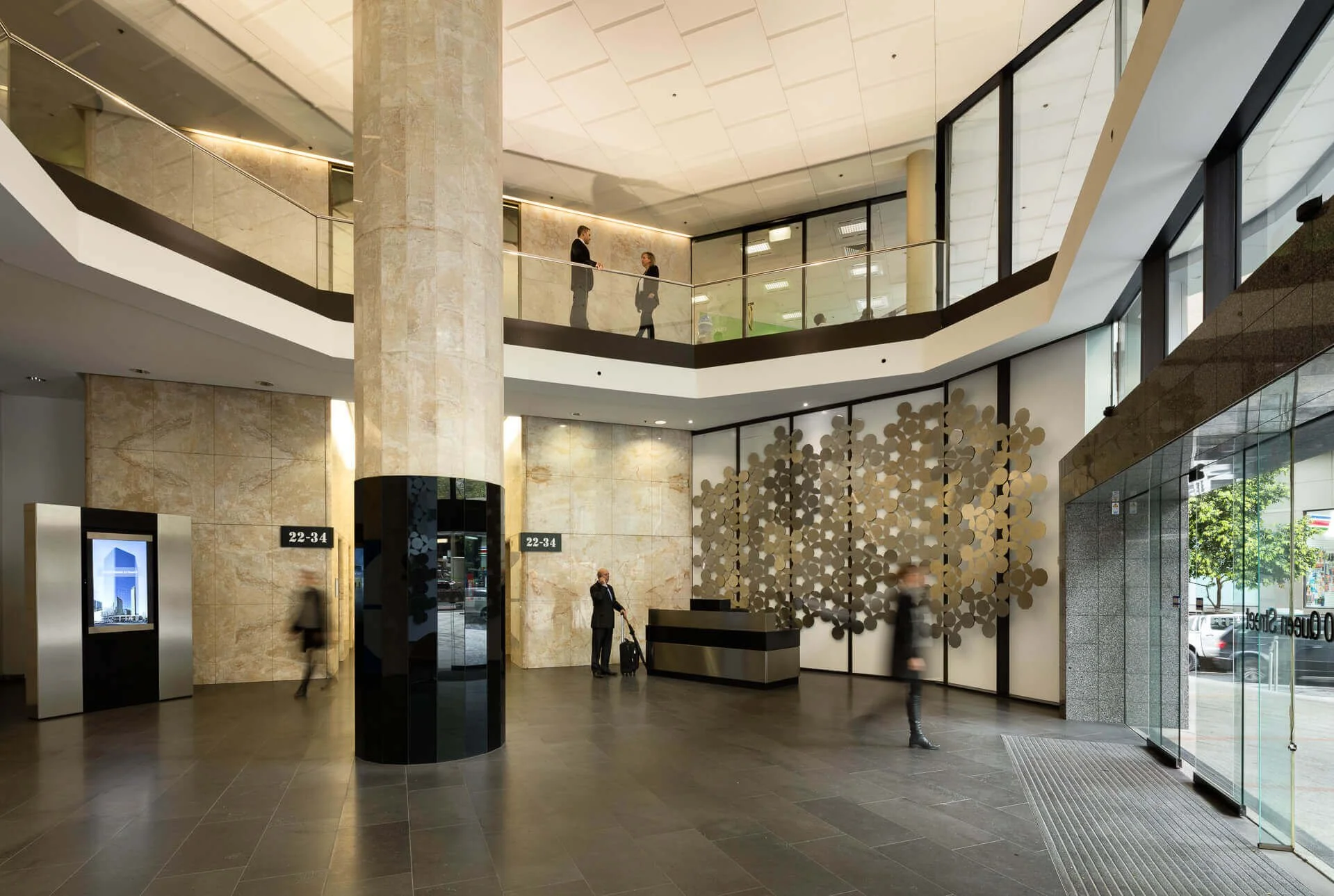 Interior of a modern office building lobby with large marble columns, glass walls, and a reception desk. People are walking and talking, and a person with a suitcase is standing near the desk.