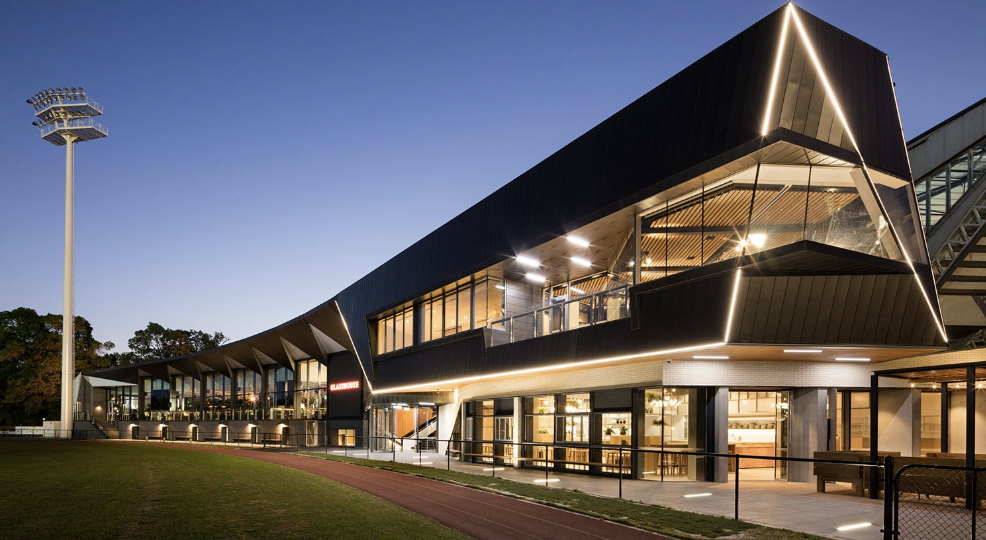 Modern building with black exterior and large glass windows, illuminated at dusk, with a sports field and tall stadium lights nearby.