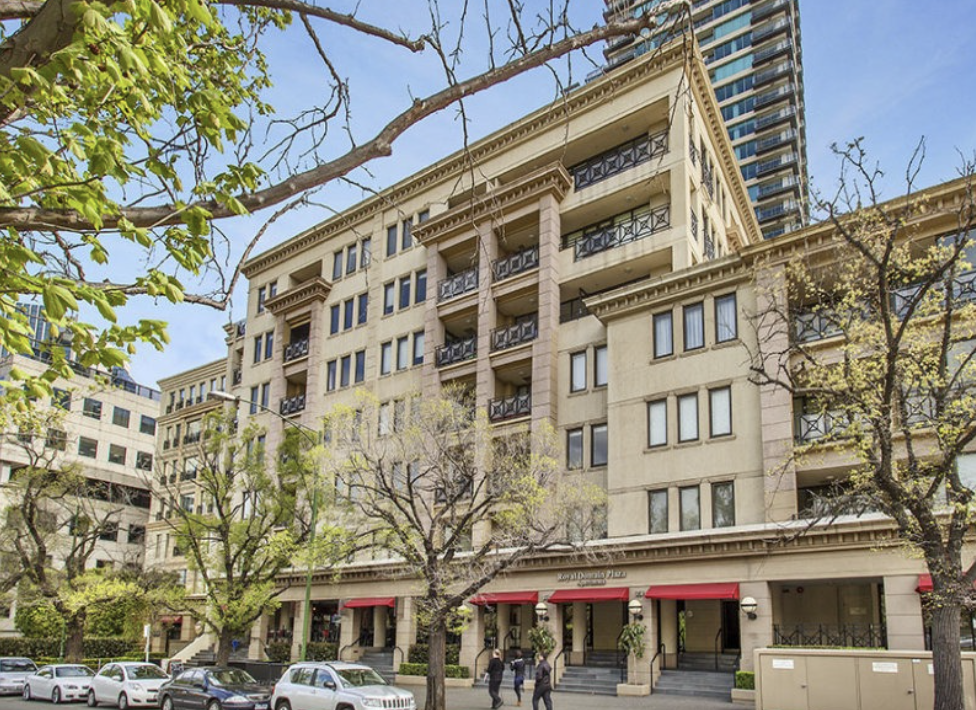 A multi-story beige building with red awnings on the ground level, trees lining the sidewalk, parked cars, and people walking in front on a sunny day.