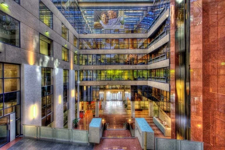 Interior view of a modern office building atrium with multiple floors, glass walls and railings, concrete and glass construction, and artwork hanging from the ceiling.