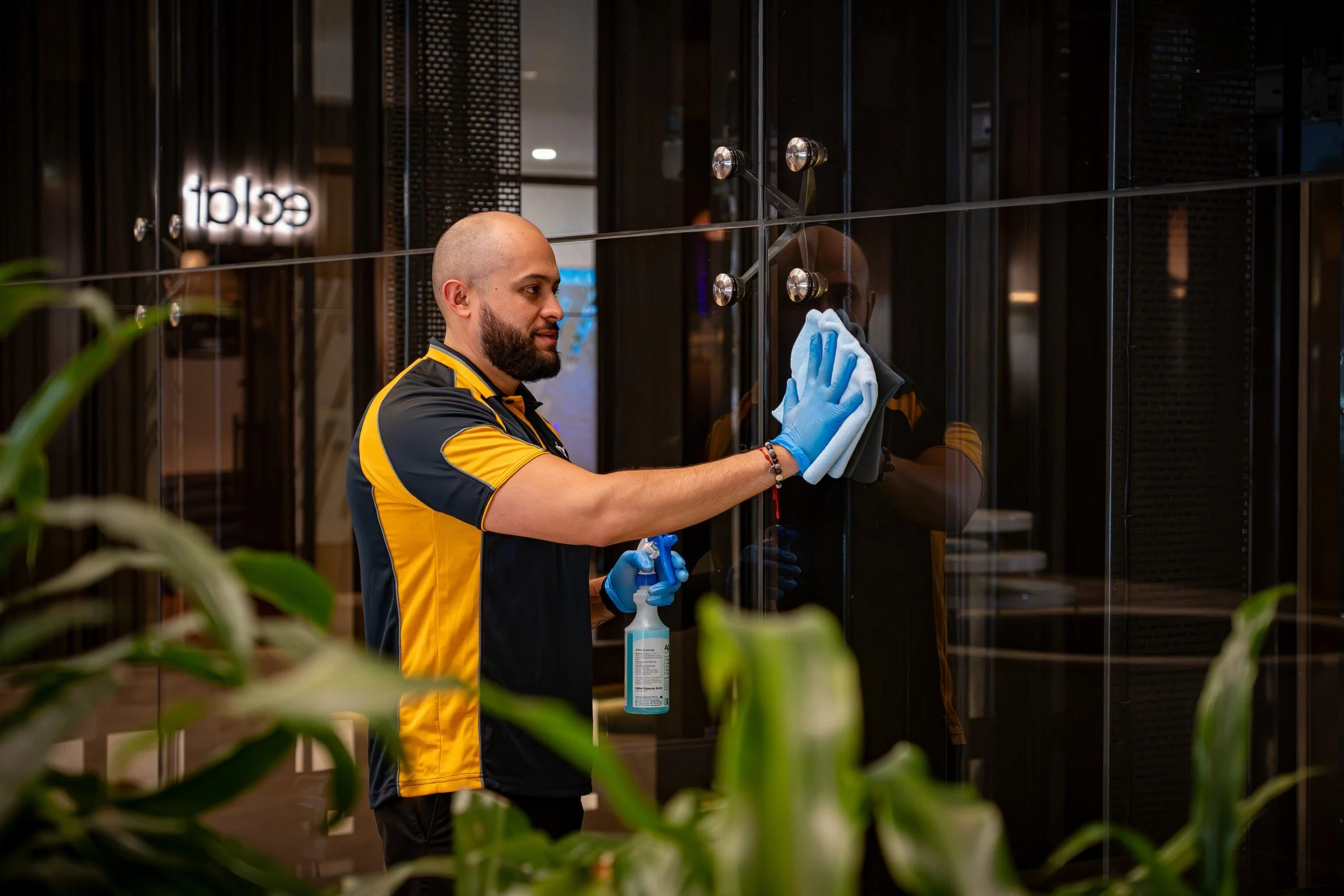 A man with a bald head and beard cleaning a glass wall or door using a spray bottle and a cloth. He is wearing a black and yellow shirt and blue gloves. The setting appears to be a modern indoor space, possibly an office or commercial building.