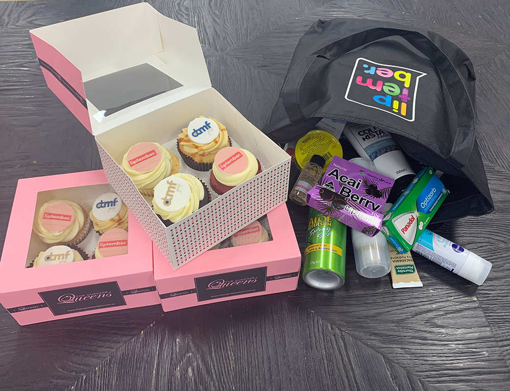 Open pink and white boxes of cupcakes with white and pink frosting, some topped with 'dmf' and 'lipember' logos, on a dark surface next to a black tote bag filled with personal care products including aloe gel, toothpaste, and medication.