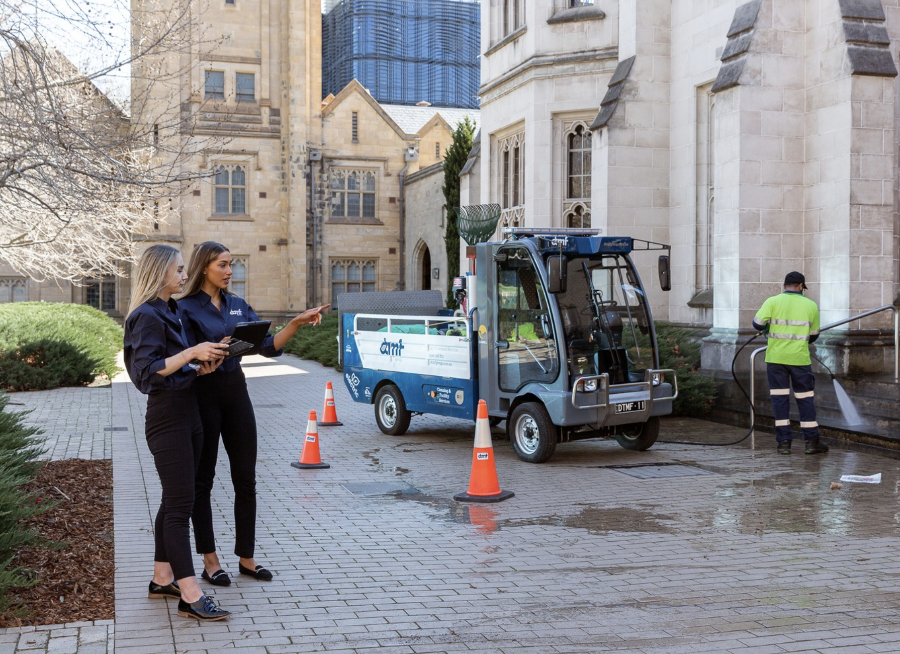 Two women in dark clothing talk while a worker in a high-visibility vest pressure washes the steps of a historic stone building. A small cleaning vehicle and traffic cones are nearby, with leafless trees and modern skyscrapers in the background.
