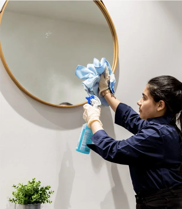 A woman cleaning a round mirror with a spray bottle and a cloth, wearing gloves.