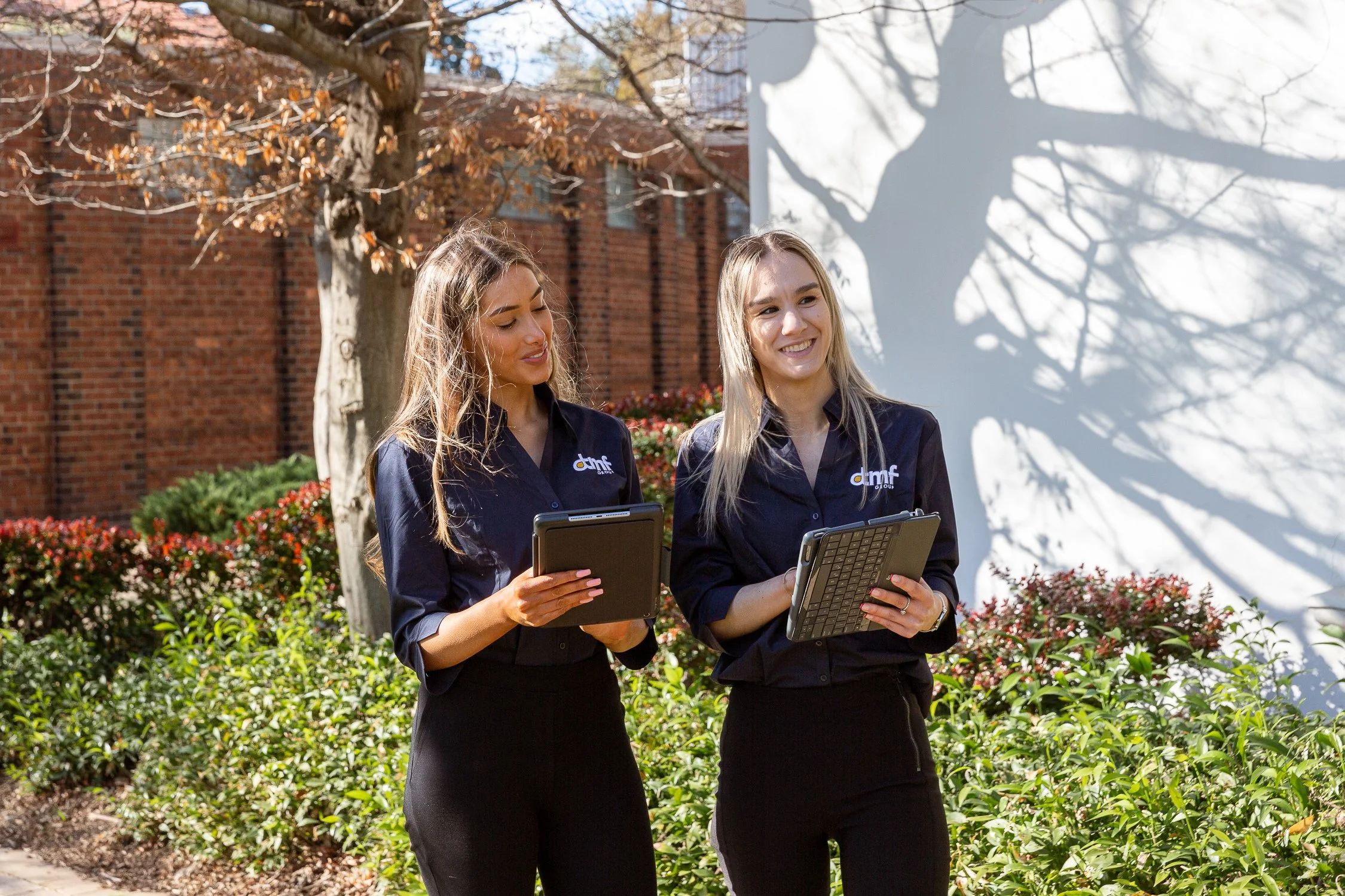 Two women standing outdoors in front of a garden and brick building, smiling and looking at tablets they are holding, with trees casting shadows on a white wall behind them.