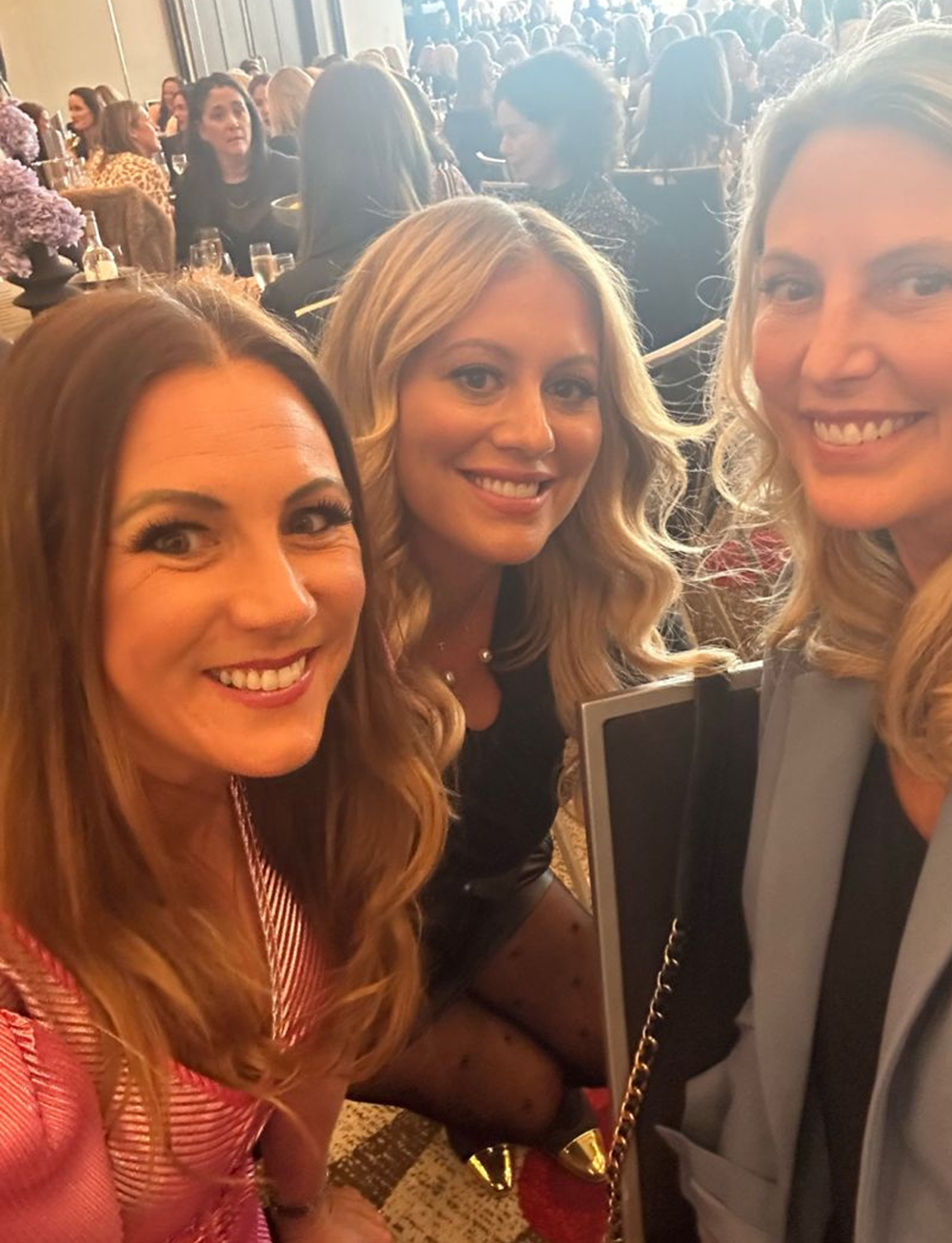Three women smiling at a social event with many other people in the background.