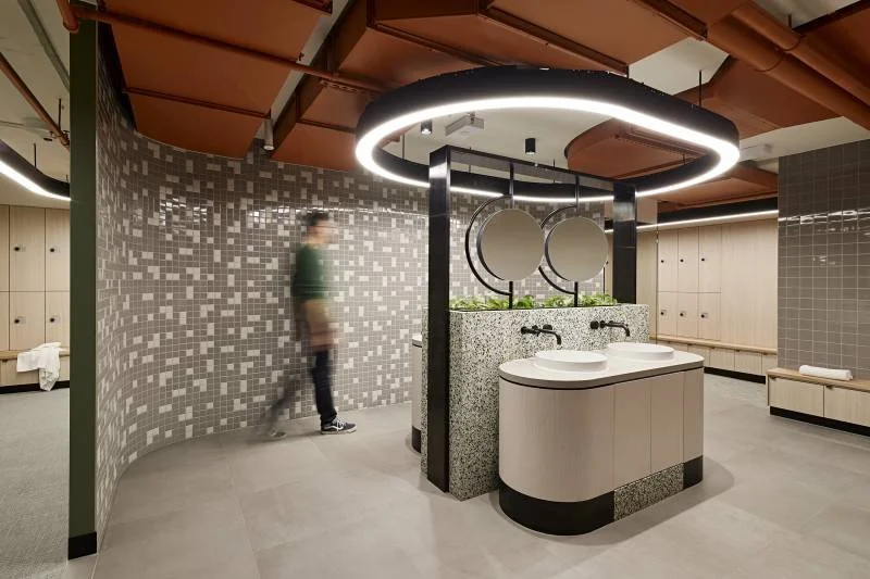 Modern locker room with a circular mirror lighting fixture and a dual sink vanity with circular mirrors, tiled walls, lockers, and a person walking past.