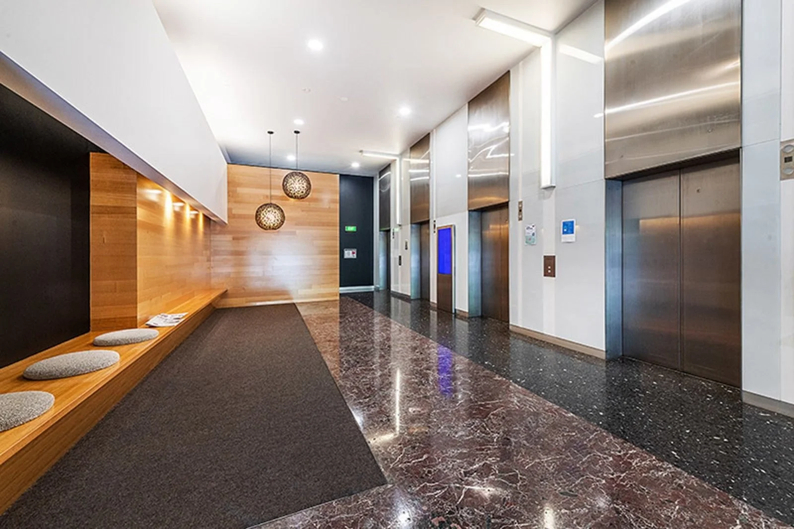 Modern building lobby with wooden wall, decorative hanging lights, three elevators, marble flooring, and sitting area with cushions