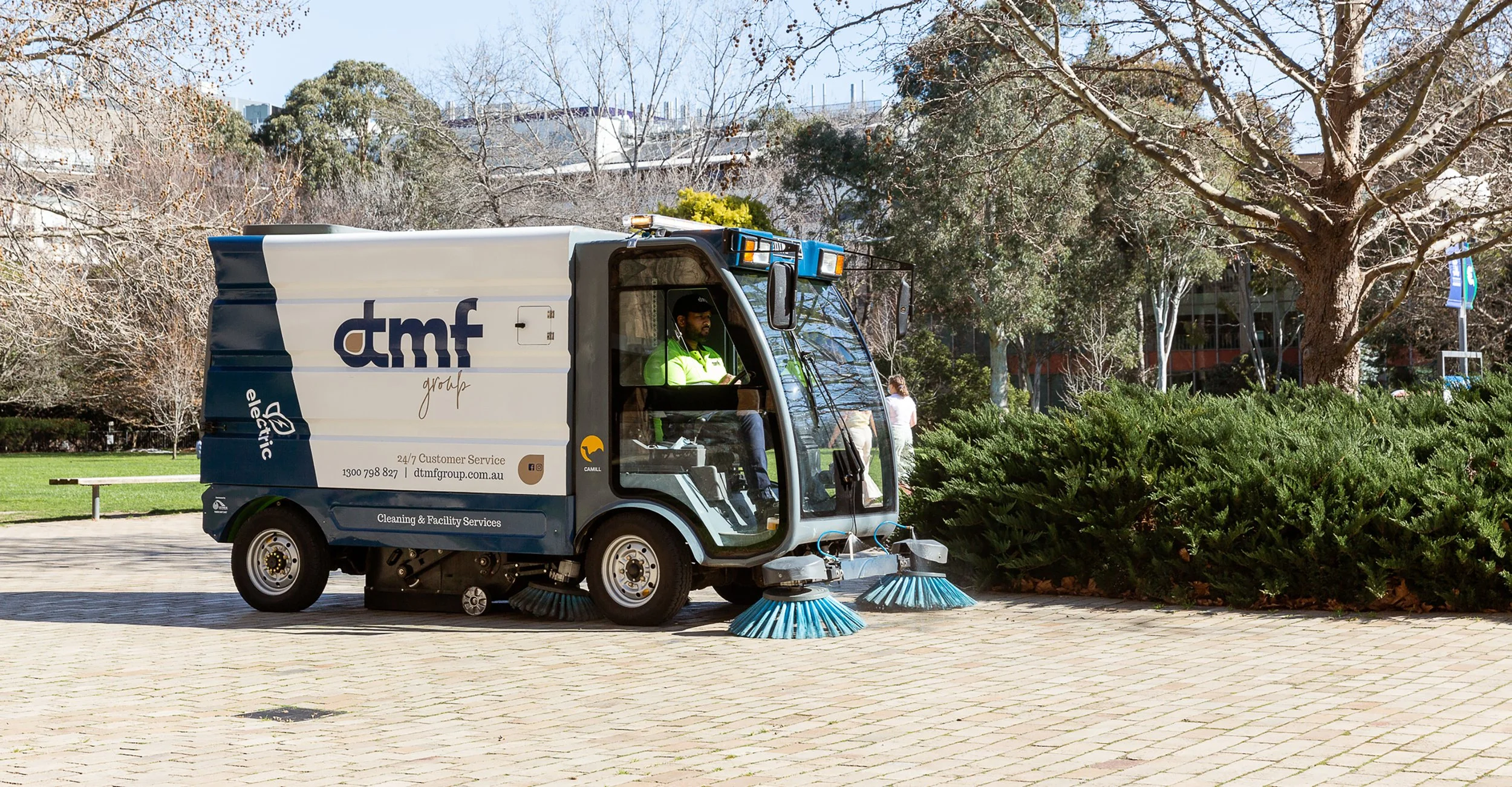 A street sweeper vehicle with a blue and white color scheme, labeled 'dtmf' and 'Cleaning & Facility Services,' operates on a paved area in a park, with trees and bushes, under a clear sky.