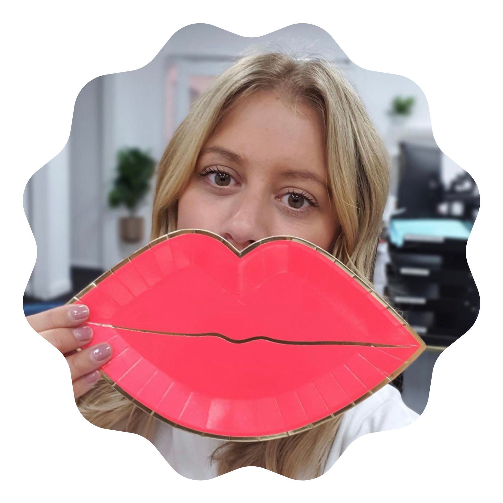 A woman with blonde hair holds a large red lip-shaped object in front of her face, inside what appears to be an office or workspace.