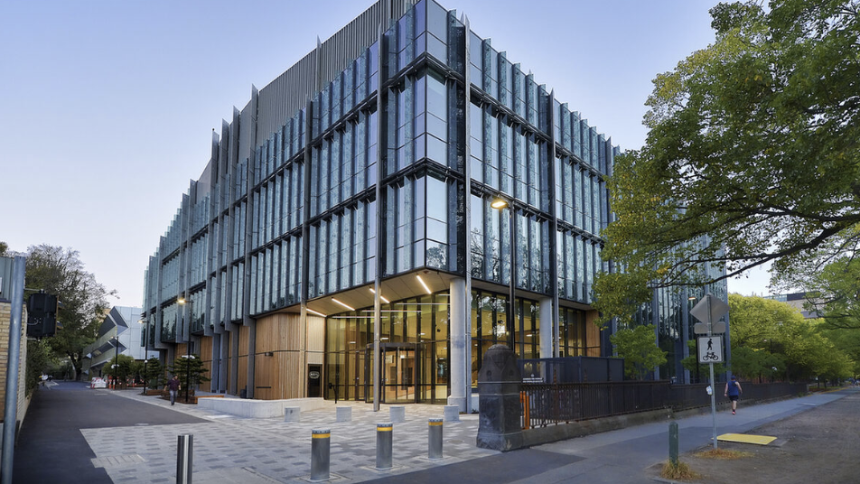 Modern multi-story glass office building on a city street with trees and pedestrians.