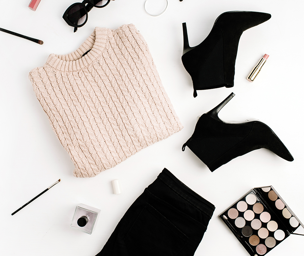 Flat lay of fashion items including a pink knit sweater, black high heel ankle boots, black pants, black sunglasses, makeup brushes, a lipstick, eyeshadow palette, and nail polish on a white background.
