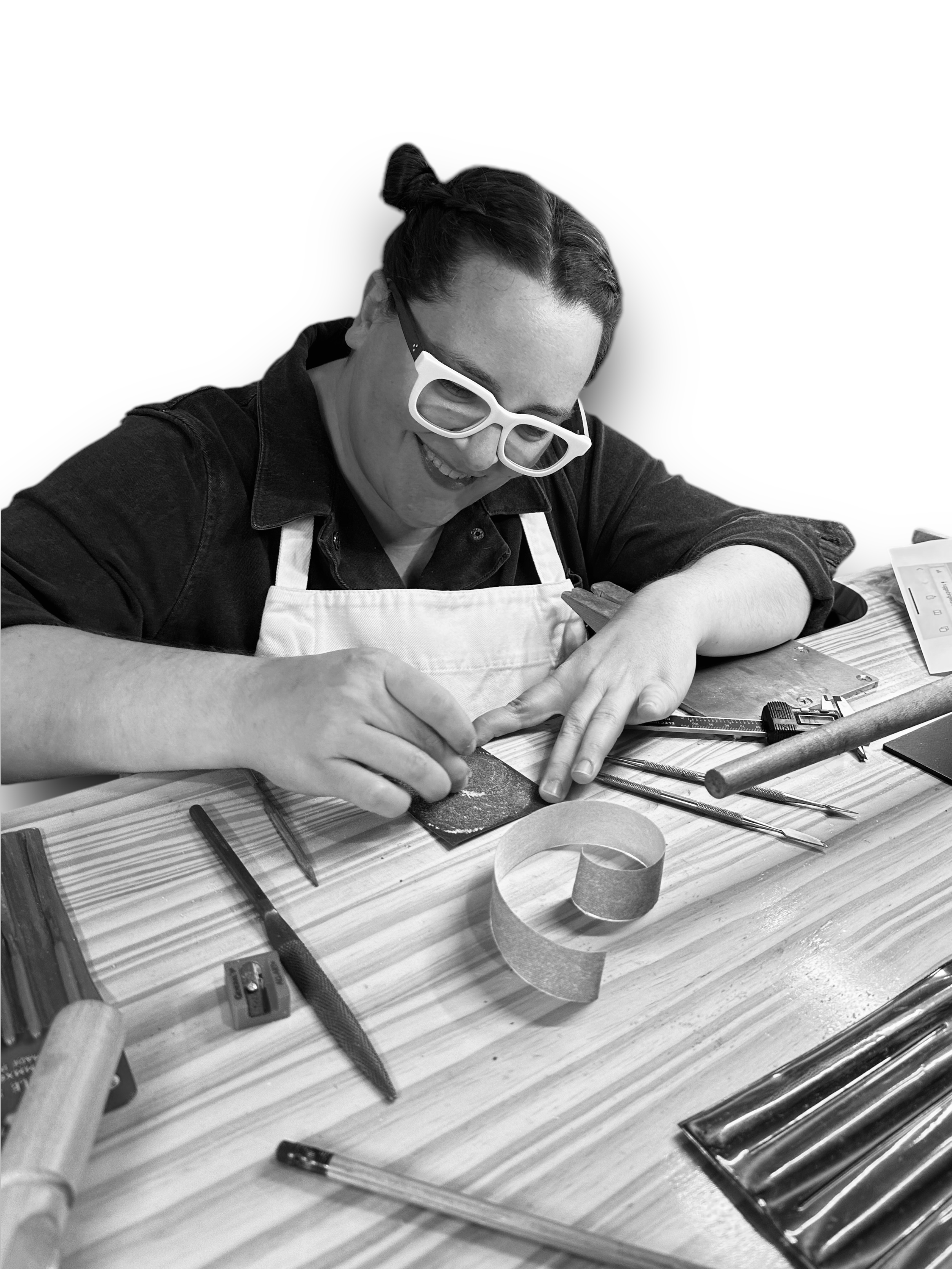 Picture of a person with pale skin wearing a black shirt working at a wood workbench. They are looking down through their glasses and smiling at the small object they are working on. They are surrounded by metalsmithing tools, pencils, and sharpeners