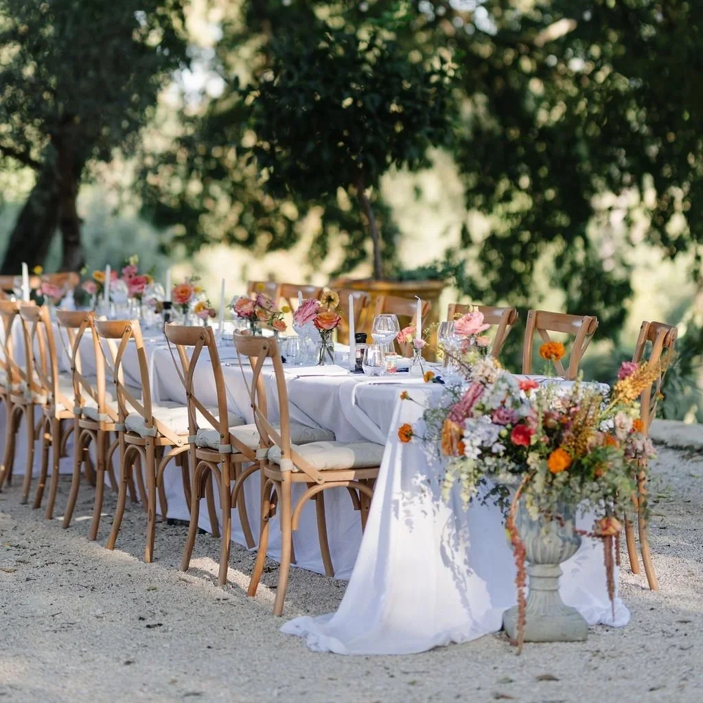 Set table with white tablecloth, beige napkin, printed menu tied with white ribbon, and silverware, with a floral centerpiece and glassware in the background.