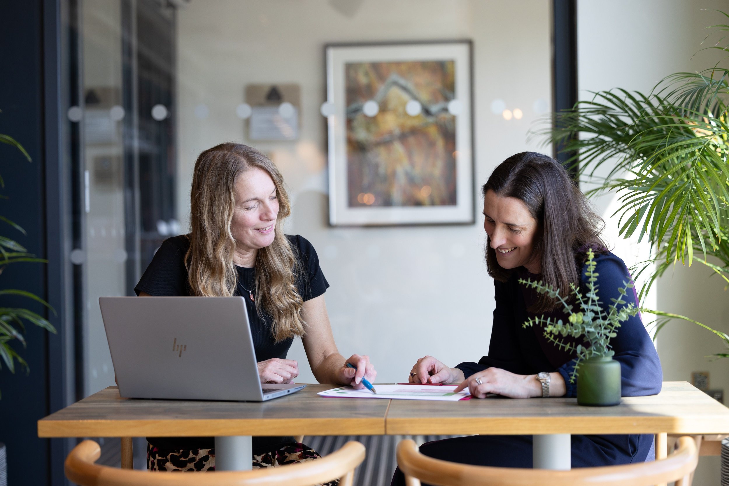 Kathy Seymour and Katy Willis sitting at a table with a laptop and papers, discussing and smiling, in a bright office setting with plants.