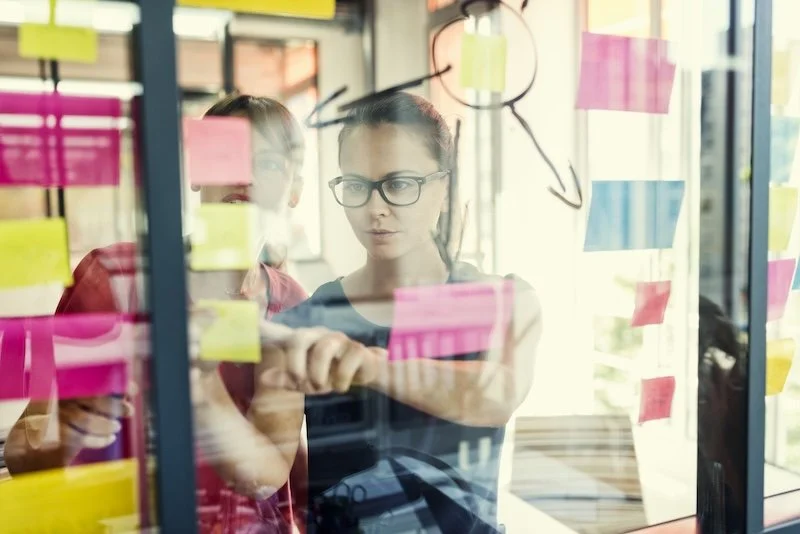 Two people brainstorming on a glass wall filled with colorful sticky notes and diagrams.