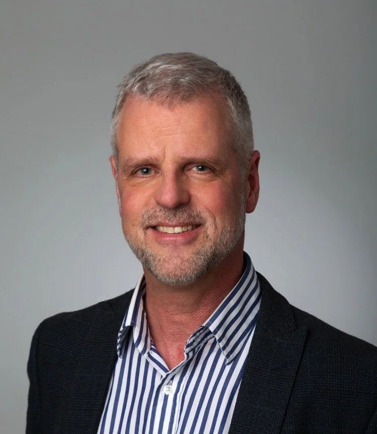 Dr Tom Loya with short gray hair and a beard, wearing a striped shirt and dark blazer, against a plain background.
