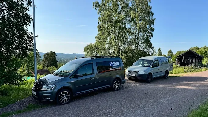 Två skåpbilar parkerade på sidan av en landsväg med gröna träd och landskap i bakgrunden.