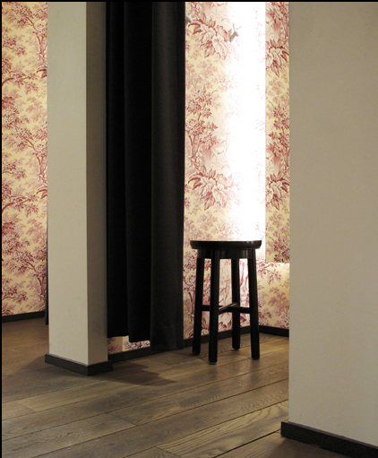 A black stool placed next to a wall with patterned wallpaper and curtains.