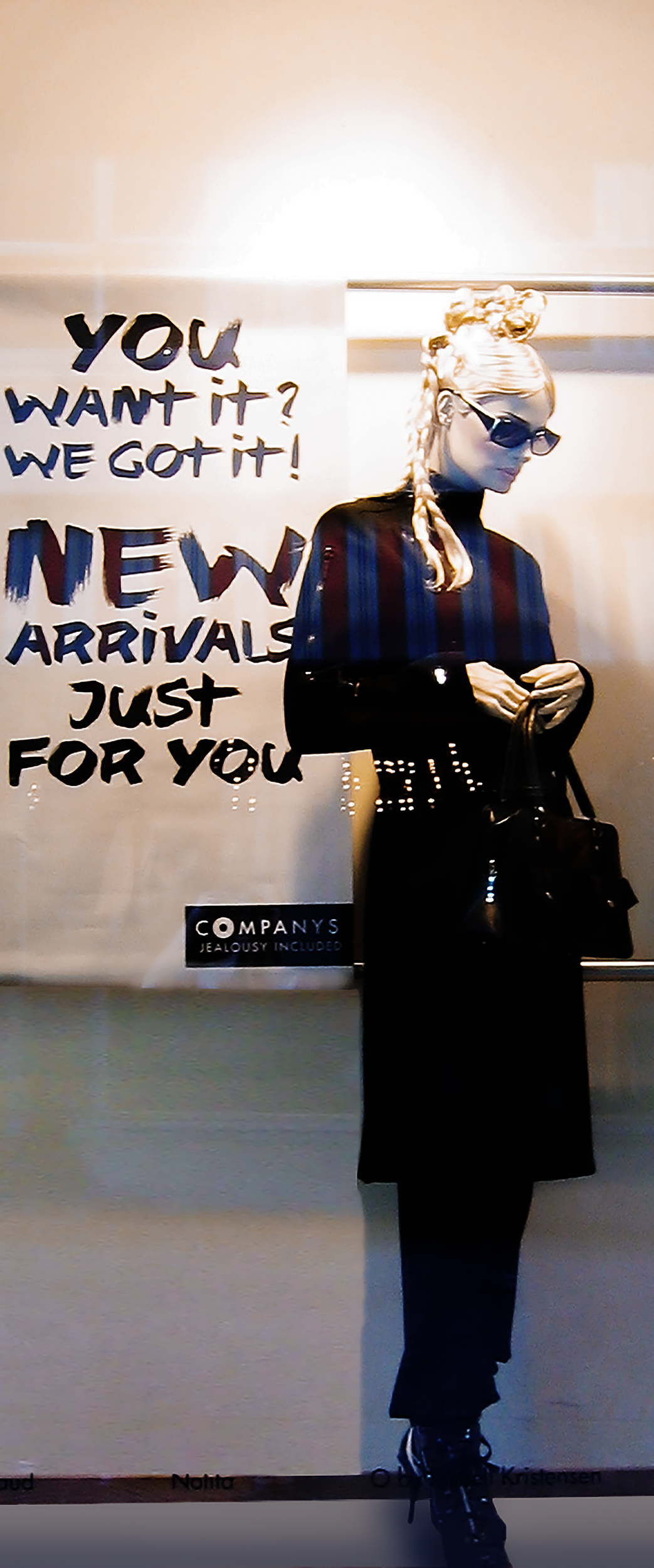 A mannequin with braided blonde hair, sunglasses, and all black clothing holding a black handbag stands in front of a sales sign at a store window. The sign reads, 'You want it? We got it! New arrivals just for you' and there is a small sign at the b