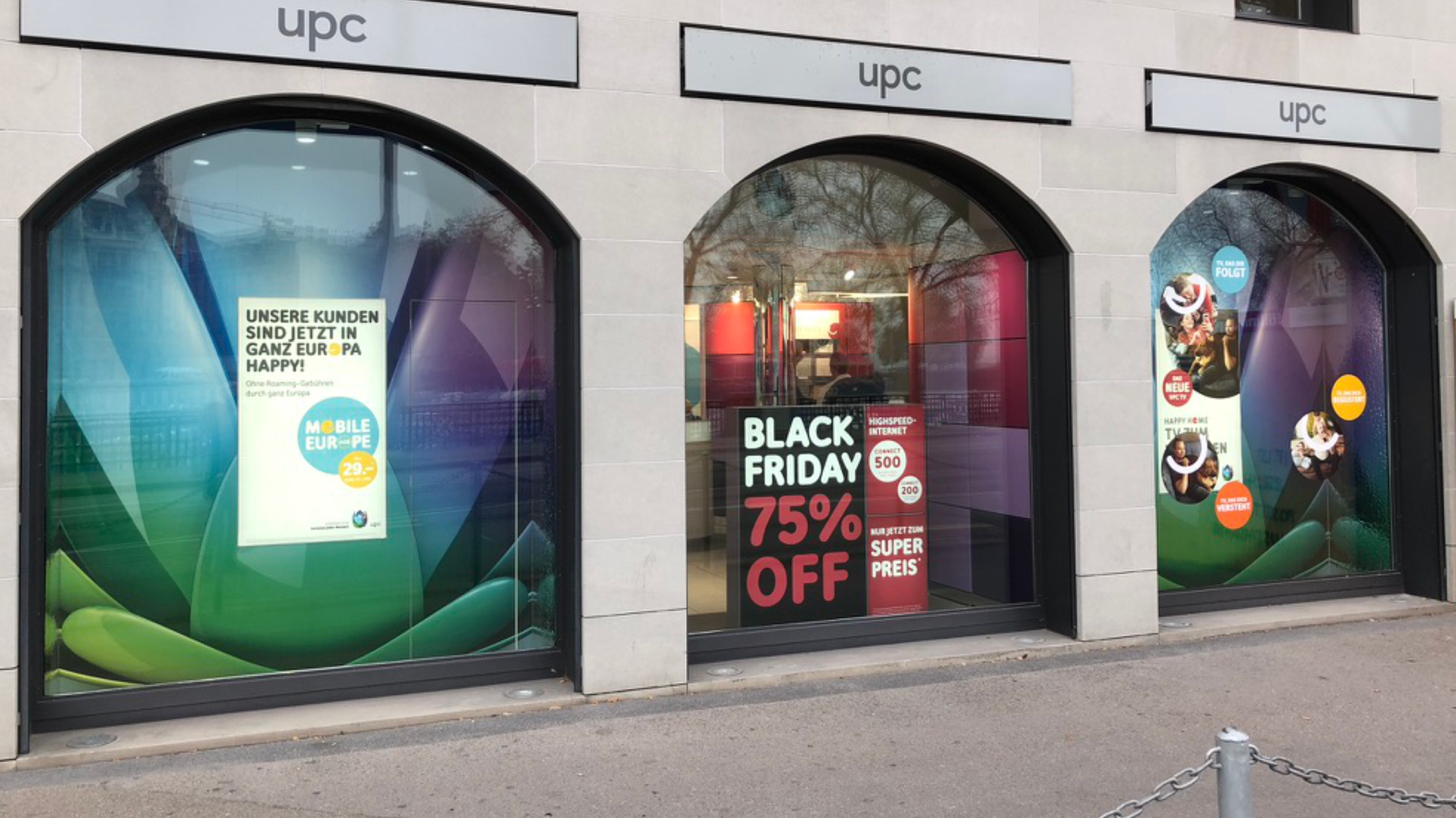Storefront with three large arched glass windows displaying advertisements, including a Black Friday sale sign offering 75% off and a promotional poster in German with customer icons and graphics, and the store logo 'UPC' above each window.