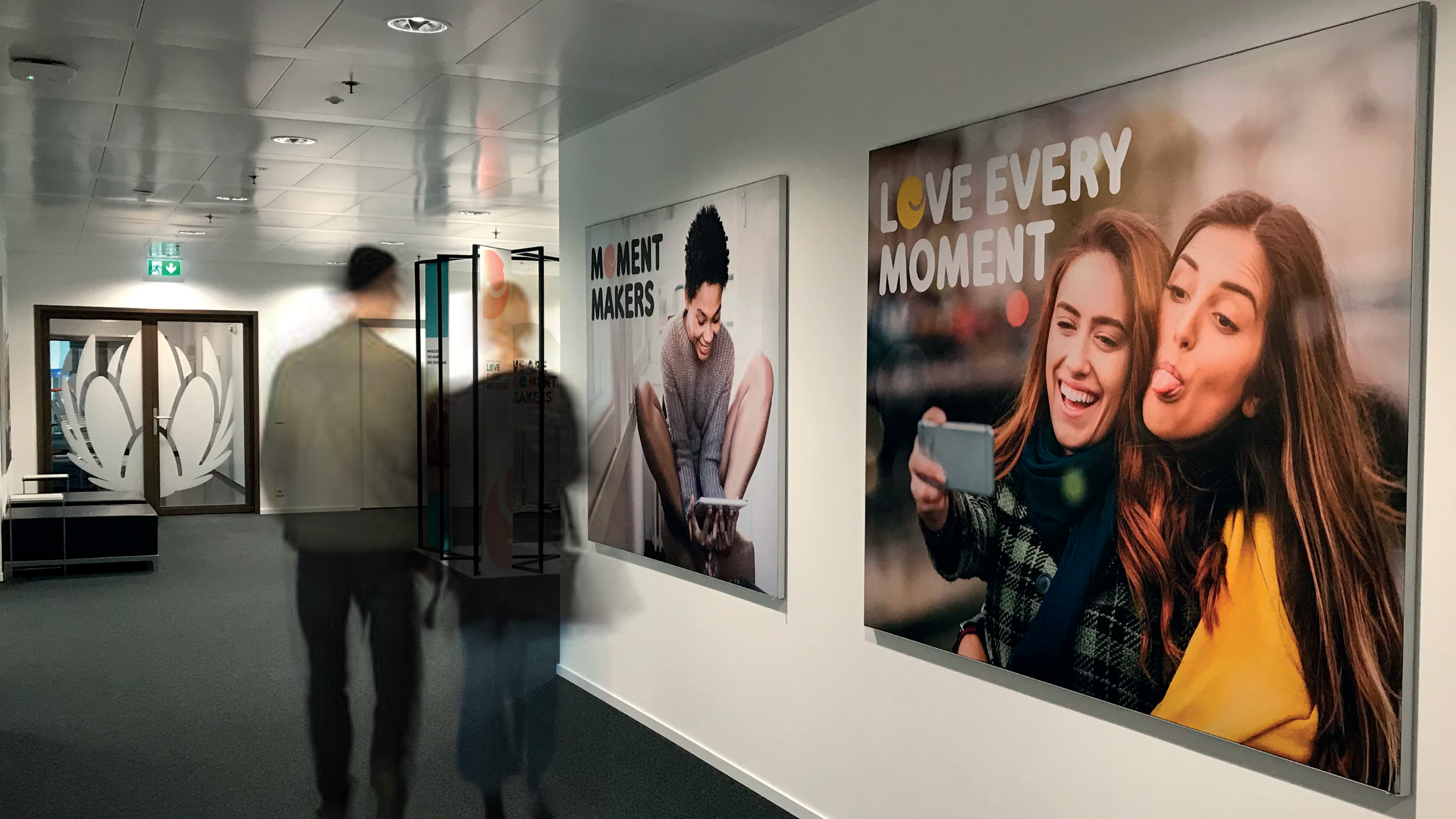 A hallway in an indoor space with two large posters on the wall featuring women using a smartphone, with positive messages about enjoying moments.