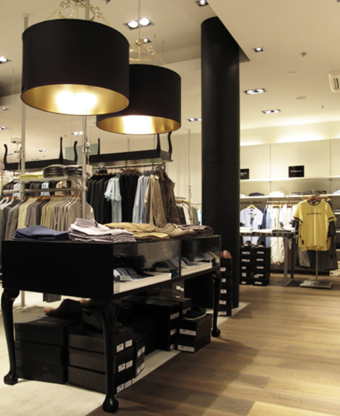 Clothing store interior with racks of shirts, jackets, and pants, black and gold ceiling lights, black display table with folded clothes and shoe boxes, wooden flooring, and white walls.