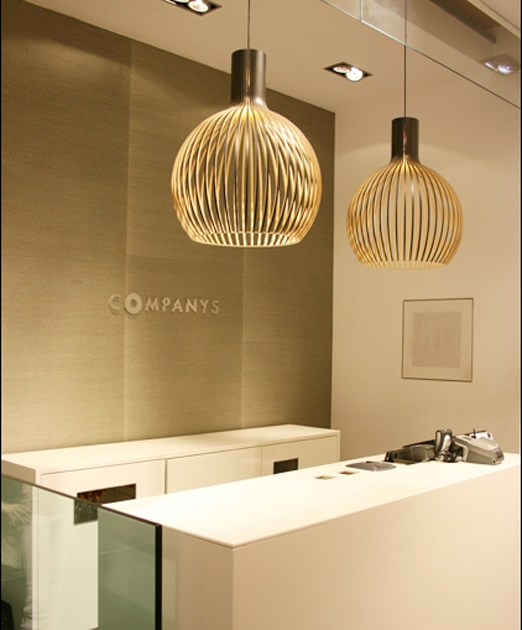 Modern office reception area with white desk, two large hanging pendant lights, and a beige wall with the word 'COMPANYS' displayed.