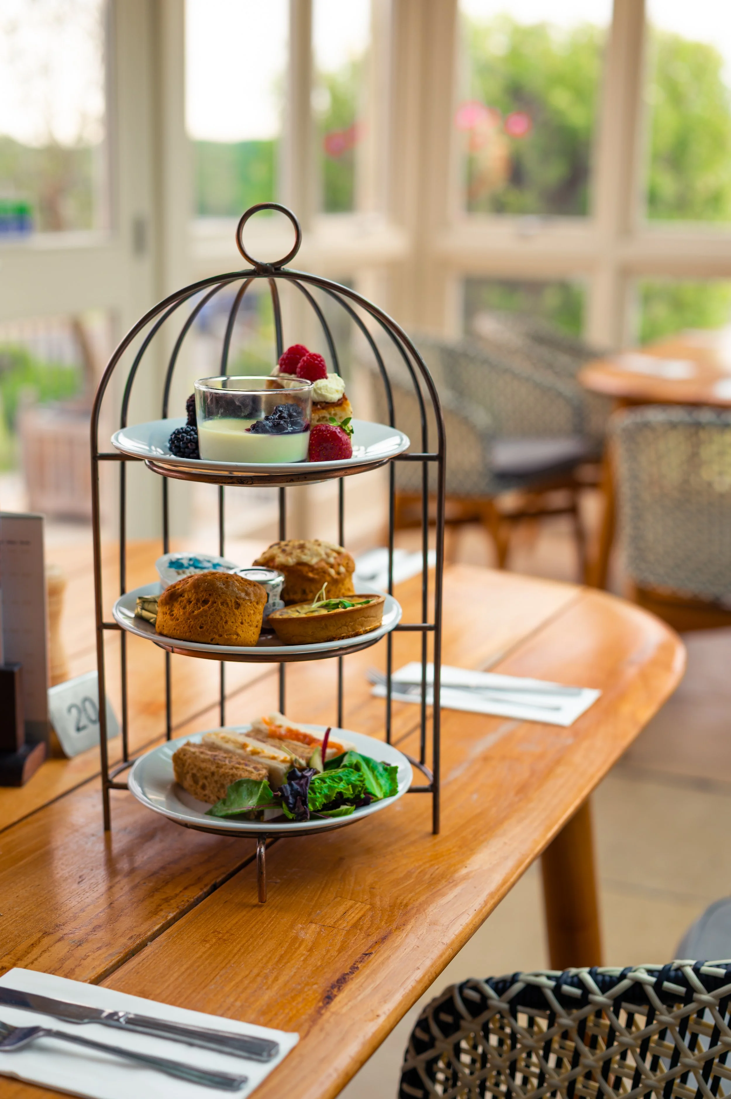 A tiered breakfast tray on a wooden table inside a bright café, holding various food items including a glass of coconut pudding, berries, muffins, sandwiches, and a salad, with chairs and large windows in the background.