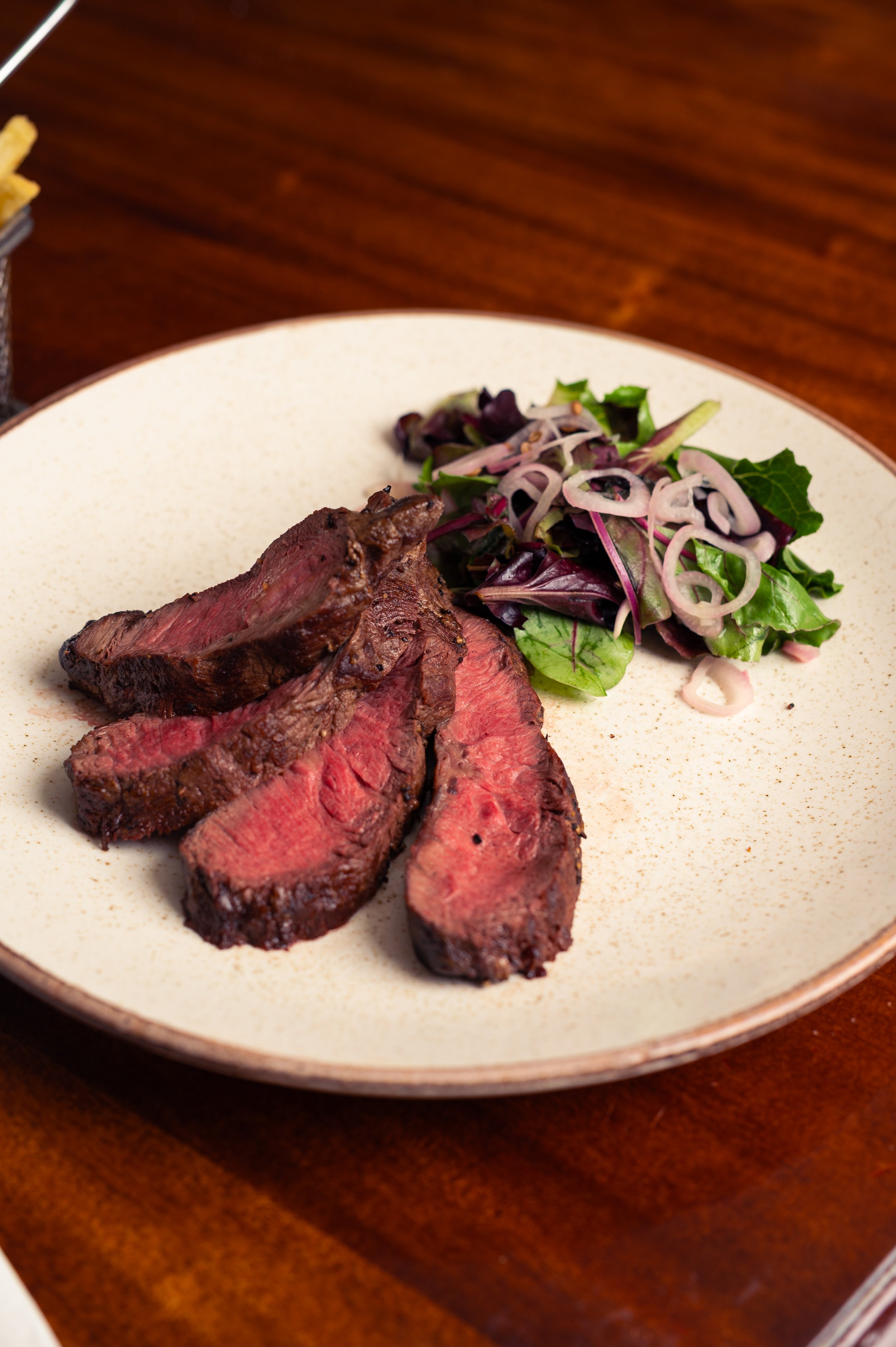 Slices of cooked beef steak with a side salad of mixed greens and onions, served on a beige plate.